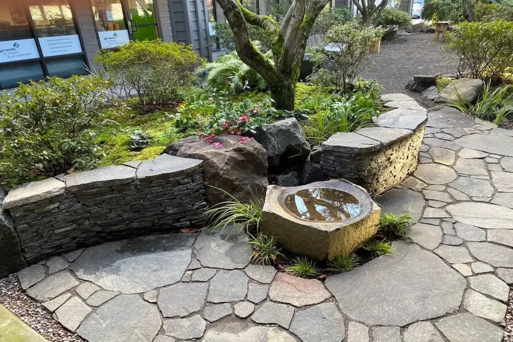 A Japanese Zen garden with an irregular flagstone pathway, a stone water feature, and architectural rock retaining walls covered in moss.
