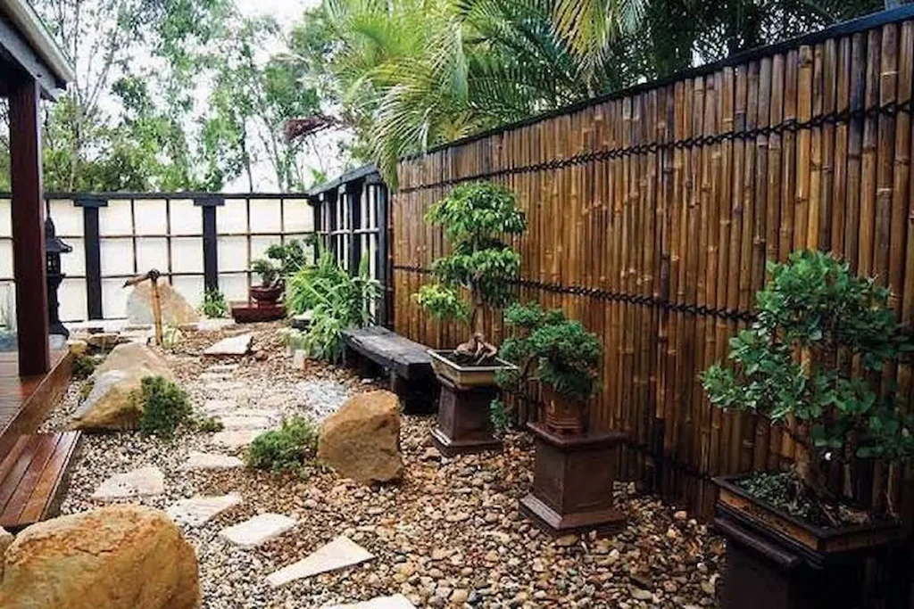 A Japanese Zen garden with a vertical natural bamboo fence, potted bonsai trees, and a stone stepping path.