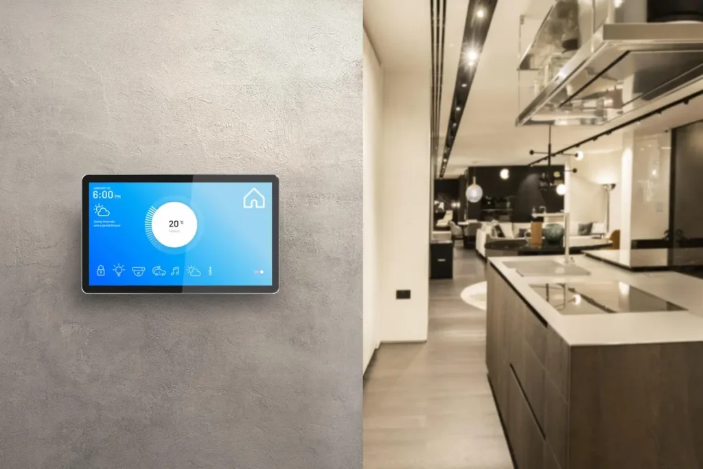 A sleek smart home control panel mounted on a concrete wall next to a modern open-concept kitchen layout.
