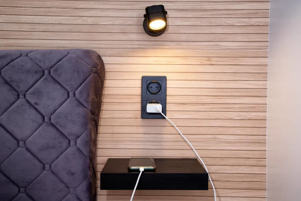 A minimalist bedside charging station with a sleek black wall outlet integrated into a light timber slat wall panel.