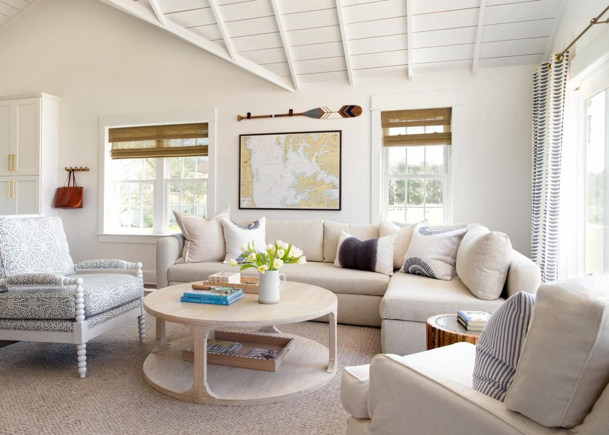 Light-filled Hamptons style living room with a neutral L-shaped sectional sofa, round wooden coffee table, and coastal wall decor