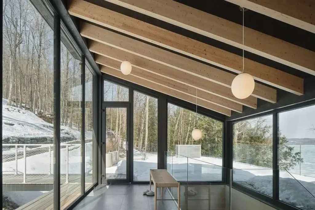 A modern geometric sunroom interior featuring heavy timber ceiling beams and dark-framed insulated thermal-break windows overlooking a snowy winter landscape.