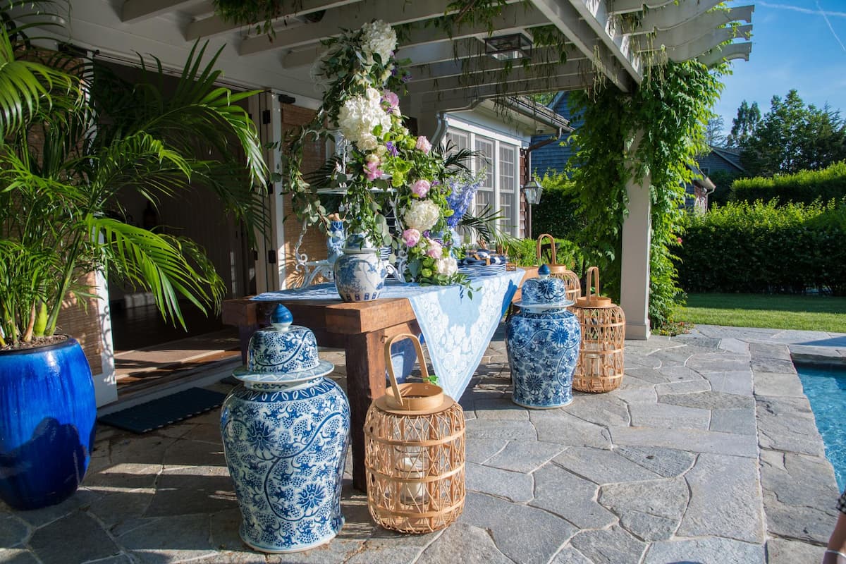 Hamptons style outdoor patio decor featuring blue and white ginger jars, lanterns, and a rustic wooden table with floral arrangements